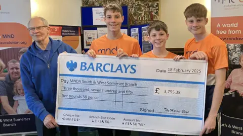 Richard Harris Three boys wearing orange tshirts present a cheque to a man in a blue jacket.