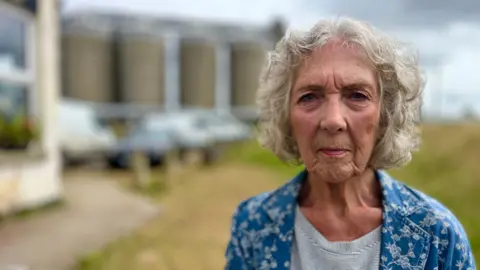 Martin Giles/BBC Dot Machin is looking at the camera and is wearing a grey top and patterned blue jacket. She has greying hair.