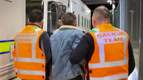 An Garda Síochána Two men in orange high-viz vests with back-up team written on the back hold a man in a denim jacket by the arms as they take him onto a police van