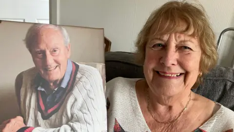 Beryl Powell, a woman with honey-coloured hair, smiles as she sits on a grey sofa holding a large photograph of her late partner, Roy Wood, an elderly man with short white hair, who is also smiling while wearing a white woollen cricket sweater and blue shirt. Beryl is wearing gold earrings, a matching necklace and a white top.