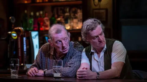 Nicola Young Photography Two middle-aged men sit at a bar, both looking at something off-camera. The man on the left has thinning grey hair and is wearing a check shirt. The other man has thicker, curly grey hair and is wearing a short-sleeved white pinstripe shirt under a green vest. Both men have their hands folded on the bar in front of them and there are partially-drunk pints of Guinness in front of them.