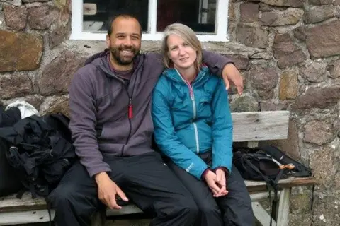 Tariq sat on the bench with Jenny, smiling. Tariq put his arm around Jenny's shoulders. Tariq has a beard and wears a purple zip-up top. Jenny has straight blond hair and wears a blue zip-up top.