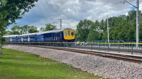 A blue train with the words Hydroflex written on the side. The front of the train is yellow with three windows. 