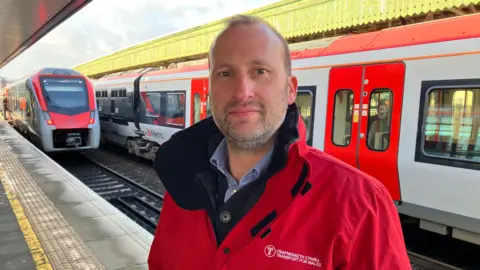 Dan Tipper works for Transport for Wales, he is wearing a red jacket and is standing on a platform with a TFW train behind him.