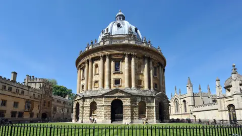 BBC Oxford's Radcliffe Camera building a sunny day - with blue skies above and green grass in the foreground. The building is dome-shaped and made from light-coloured stone. There are several other buildings in the background made from the same stone.