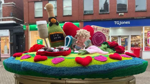 Knitted representations of a Champagne flute, Champagne bottle, box of chocolates, Cupid figure, a heart shape and a Valentines card. They are sat on top of a gold post box. There is an opticians and newsagents shop in the background.