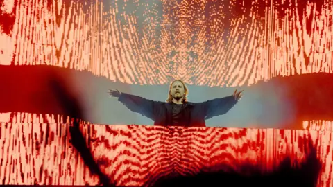 Elliot Hingston A man performing in dark clothing, with a bright orange staging effect around him. 