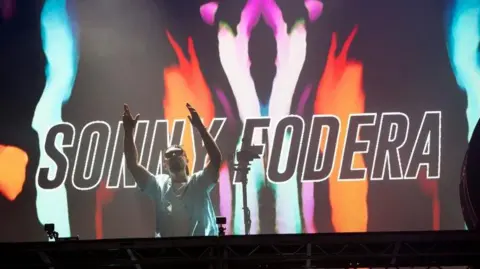 DJ Sonny Fodera on stage in front of a colourful backdrop featuring his name. Sonny has his arms aloft and is wearing sunglasses