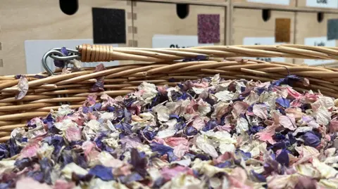 Ellen Knight/BBC A close-up photograph of a large basket filled with dried petals, a mix of purple, pink and white. Behind the basket is a row of wooden boxes. 
