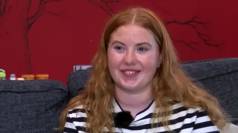 BBC A young girl with ginger hair and a black and white horizontally-striped top smiles while sitting on a couch