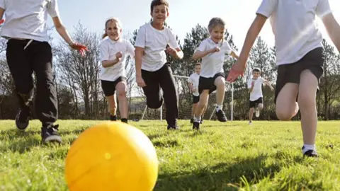 Children run towards a yellow ball as they play on the grass
