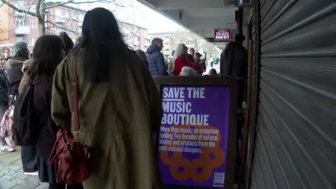 A group of people queue outside a shop front with a purple "Save The Music Boutique" sign propped outside.
