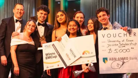 Alexander Horswill A group of smartly-dressed students smiling with three large cards that indicate their victory and their prize money