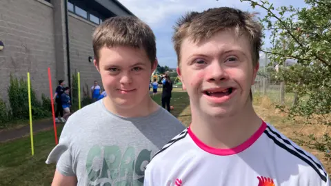 Peter and Aren are standing together. One wears a white O'Neill top with a pink collar and black stripes running down the shoulders. The other is wearing a grey t-shirt with faded green lettering on the front. 