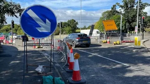Traffic passing roadworks controlled by temporary barriers and cones at a road junction, with a road closure sign visible in the foreground.