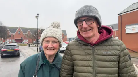 A man and a woman smiling at the camera. The woman, on the left, is wearing a cream bobble hat and a turquoise coat. The man, on the right, is wearing a green puffer jacket and a grey hat. They are standing in front of a car park and modern rows of shops.