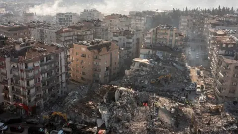 Reuters An aerial image of a destroyed city in Turkey following the 2023 earthquakes. It shows collapsed and burning tower buildings as far as the eye can see. In the foreground there is a huge mound of rubble where a building once stood. White smoke is rising in the distance. White vans and heavy machinery can be seen on the roads, searching for survivors and clearing rubble.