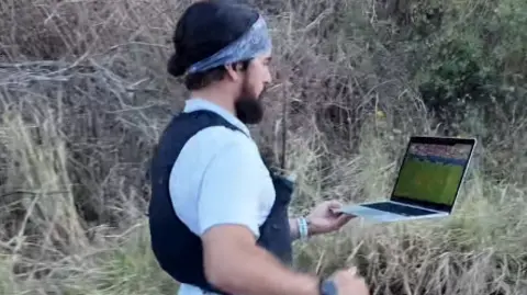 Mike Humphreys/Juan Cruz Lopez A man wearing black running shorts, a white T-shirt, a black running vest and a bandana. He is running on a road while holding a laptop which has a football game playing on the screen.