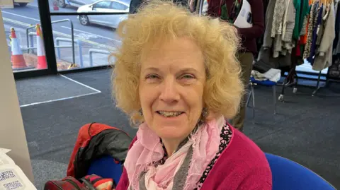 A woman with blonde hair wearing a pink cardigan and scarf. She's sat on a blue chair with a window behind her.