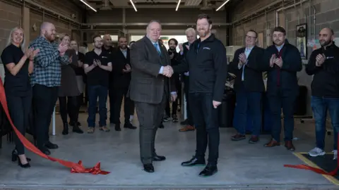 Goldilock Council leader Stephen Simkins and Stephen Kines shake hands after a red ribbon is cut at the expanded premises. Behind them is a crowd of about a dozen men and women clapping.