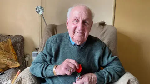 Contributed Mr Cracknell holds his Legion d'Honneur medal as his sits in a chair. He is smiling at the camera.