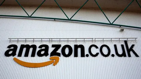 A logo sits on a wall outside the Amazon Marston Gate fulfilment centre, the building is white with green metal above. 