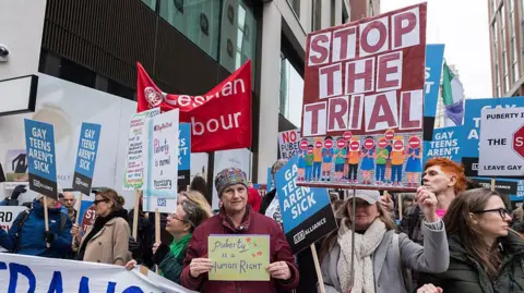 Getty Images Demonstrators pictured in central London in December 2025 protesting against the clinical trial. One sign reads "Stop the trial" and another reads "puberty is a human right". 