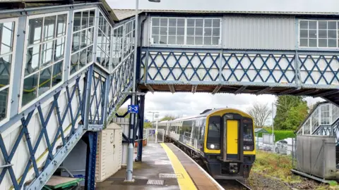 SWR A yellow fronted train approaching the platform of a station driving under a stepped footbridge painted blue and white