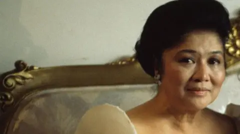 Imelda Marcos in later years with heavily lacquered dark hair and a dress with a Gomesi shoulder pad.
