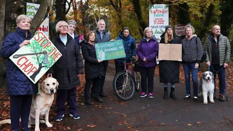 A group of residents, some holding placards saying 'Stop the chop' and 'Shorrocks Hill belongs to nature' stand on the site of the planned development. One of the residents has a golden labrador on a lead and another has a medium-sized dog next to them.