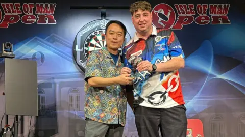 Isle of Man Darts Festival Reece on stage posing with his winners trophy. He has a colourful darts-themed polo and has scruffy brown hair.