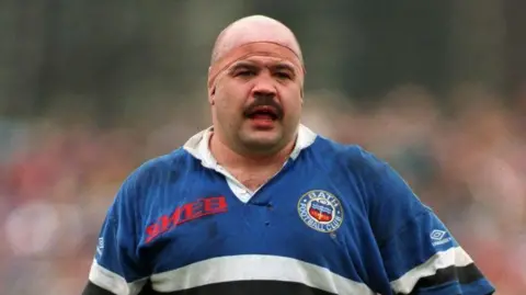 Gareth Chilcott in his rugby uniform in 1993. He has a headband around his head and a mustache.