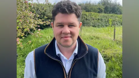 Conservatives A man with dark hair smiles at the camera, wearing a dark blue gilet and a light blue shirt, in a field next to hedgerow