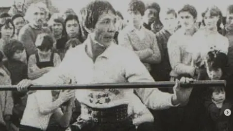 A black and white photo of a man holding a long stick in front of a crowd. 