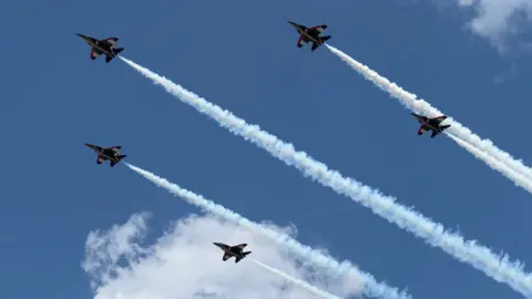 BBC Five jets, in formation, cross the largely blue sky above Farnborough, with smoke streaming behind them