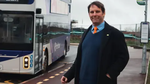 Somerset Council Richard Wilkins is standing at a bus stop as a bus is stopped next to him. He is wearing a dark blue jumper, with a light blue shirt and orange tie. He looks to be in his forties and has dark brown hair