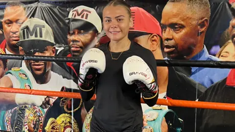 Tiah stood in front of a poster with multiple boxers behind her, she is wearing a black-long sleeved top and black shorts. She is wearing white boxing gloves. 