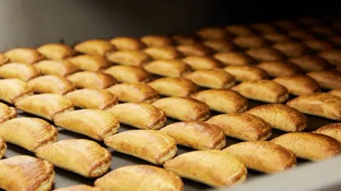 Ginsters A picture of hundreds of pasties on a conveyor belt. They are golden brown in colour.