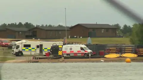 Cheshire Police A lake in the foreground with a police mobile incident unit on the shore, a search and rescue team van next to it, two crates full of canoes also on the shore, and chalet buildings in the background.
