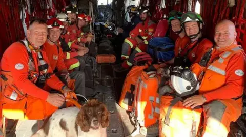 A group shot, including Mark (pictured front left, near a spaniel looking dog). This is from a UK ISAR earthquake deployment in 2023, in Morocco. The group are wearing orange overalls and are in a plane. Some are wearing helmets.