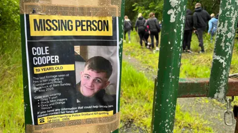 BBC Missing person poster on a gate as people seen walking behind