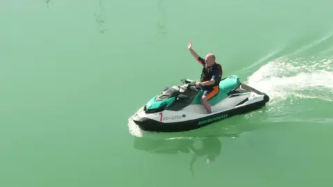 Ed Davey om a jet ski on still green water 