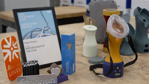 The image shows a number of awards sat on a wooden table. There is a frame which says 'New Designers 2026 Rising Star Award'. There is a orange award, a Cadbury award and Joseph Joseph award. In the background, there are a number of grey prototypes and the finished orange and blue product.