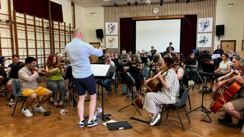 An orchestra rehearsing in a large hall, with a conductor leading them.