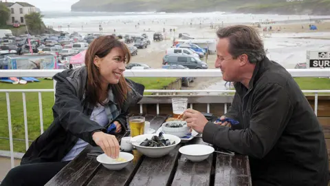 Getty Images Samantha Cameron and David Cameron sitting opposite each other on a bench at a restaurant in Polzeath, Cornwall. They are drinking and eating mussels. They are smiling at each other. In the background is Polzeath beach and a car park.