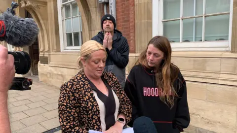 Det Con Katie Smith wearing a leopard print shirt standing next to Tracey Moore in a Hoodrich jumper with a man in the background in a black and grey coat and a black woolly hat. A TV camera is to the left of the shot. 