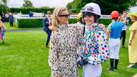 Goodwood Racecourse Two women stood hugging. One is wearing a dress and looking at the other, while the other is looking slightly towards the camera and wearing a jockey outfit. They are stood on grass. 