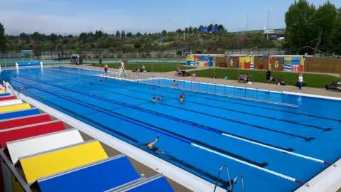 BBC Swimming pool lanes in a lido with colourful changing huts at the bottom of the picture.