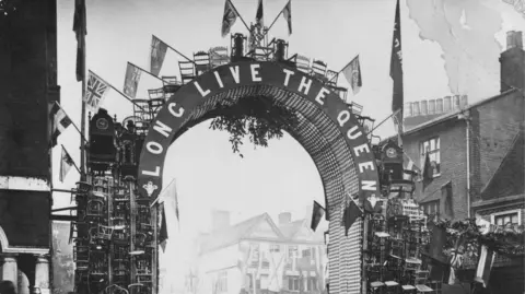 Wycombe Museum A black and white photo with an arch saying Long Live the Queen, decorated with flags and wooden chairs, in a high street