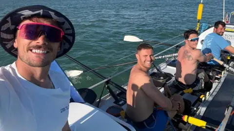 A selfie taken by a man wearing a white top, sunglasses and a hat. Behind him are three men sitting and rowing, but turning around to smile at the camera. Two are shirtless and the one sitting towards the front is wearing a blue top.